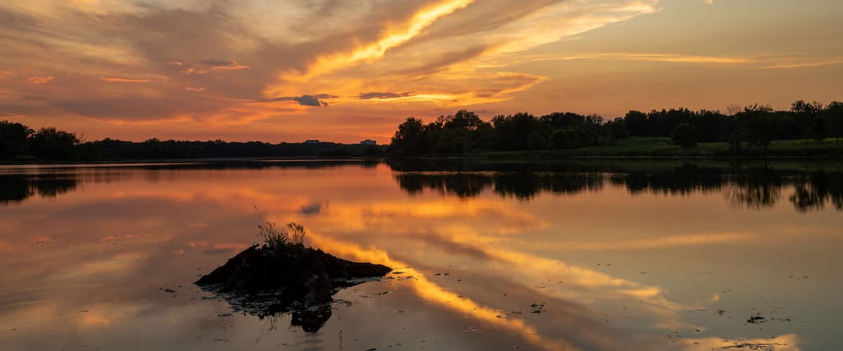 A sunset over a calm lake with a silhouette of trees and a rocky outcrop in the foreground.