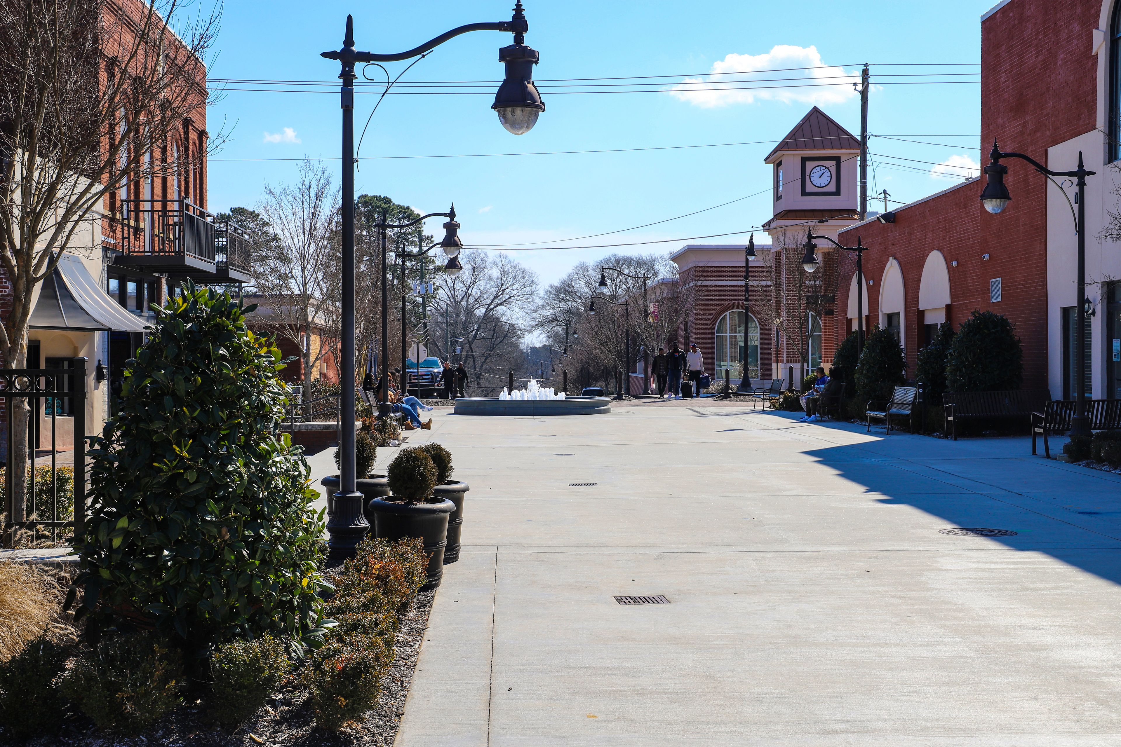 A sunny street lined with brick buildings, a clock tower, and a fountain.