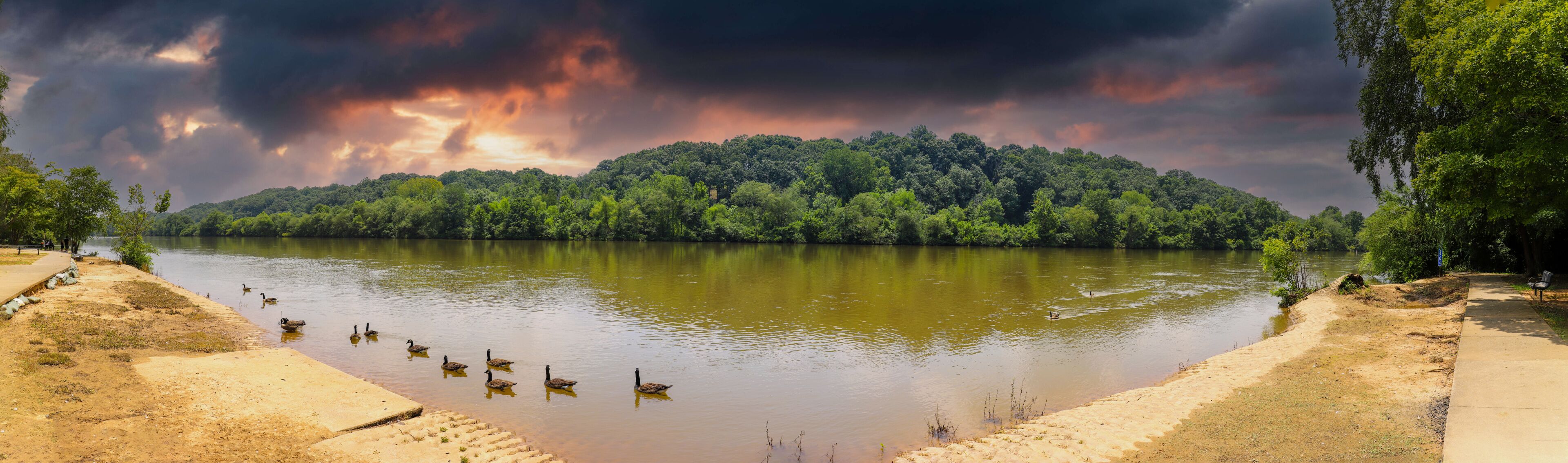 a stunning panoramic shot of the silky brown water of the Chattahoochee river surrounded by lush green trees reflecting off the water with powerful clouds at sunset at Azalea Park in Roswell Georgia