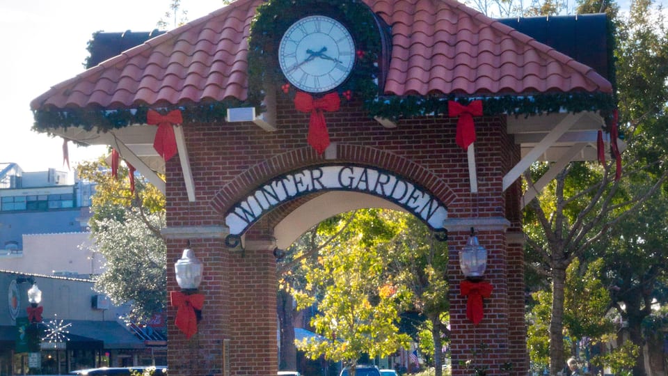 A brick archway with a clock and 'Winter Garden' sign, surrounded by greenery and a clear sky.