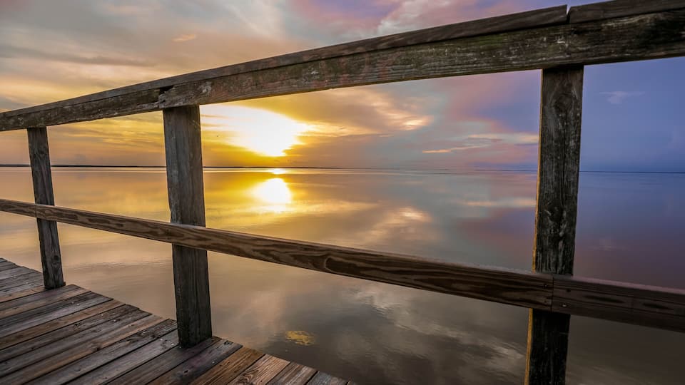 A wooden pier extending over calm water, reflecting a sunset sky.