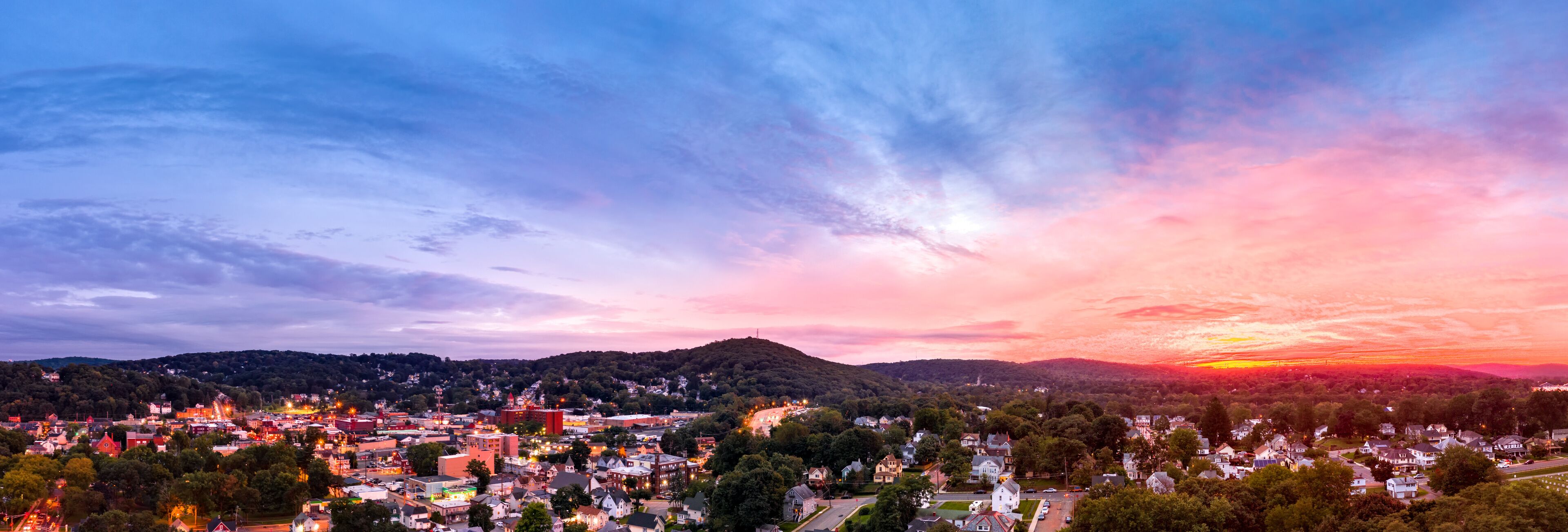 A town at sunset with a colorful sky, rolling hills, and a mix of residential and commercial buildings.