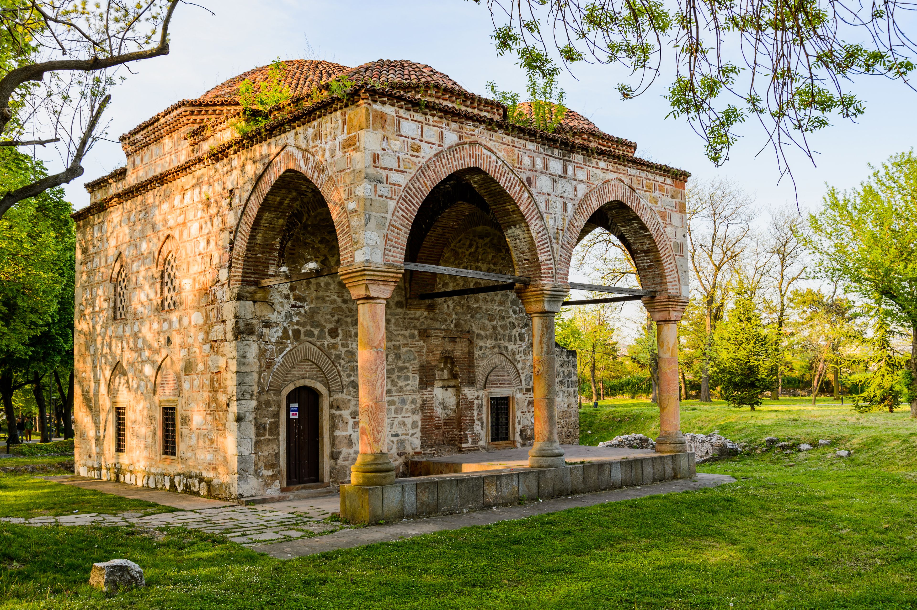 A stone structure with arches and a tiled roof, surrounded by grass and trees.
