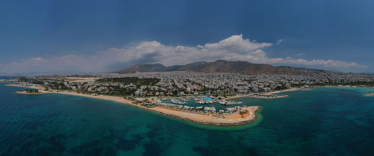 An aerial view of a coastal city with a mountainous backdrop, a marina, and clear turquoise waters.