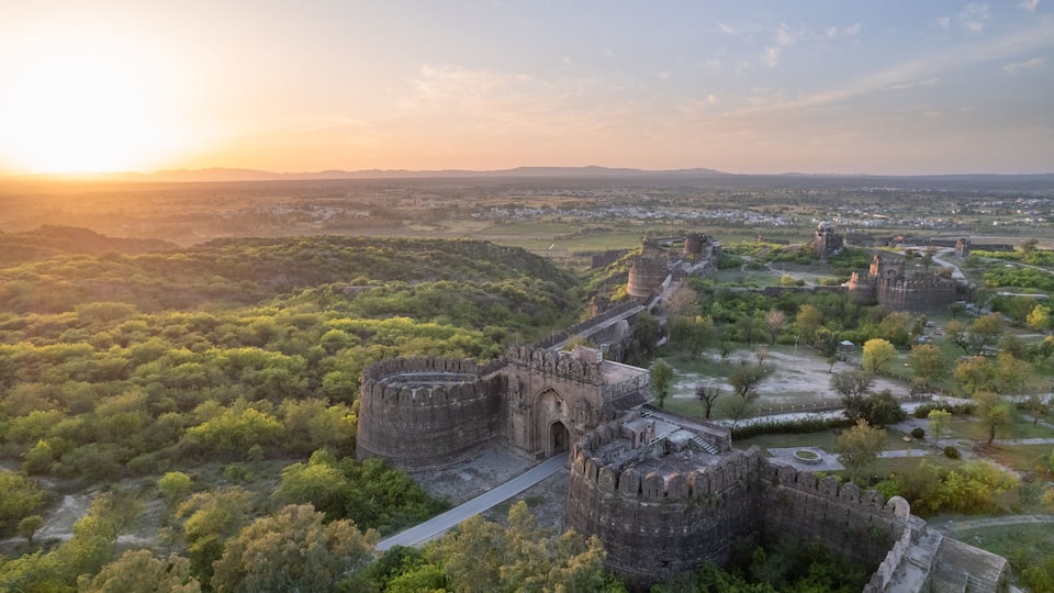 An ancient fort surrounded by lush greenery and a distant cityscape at sunset.