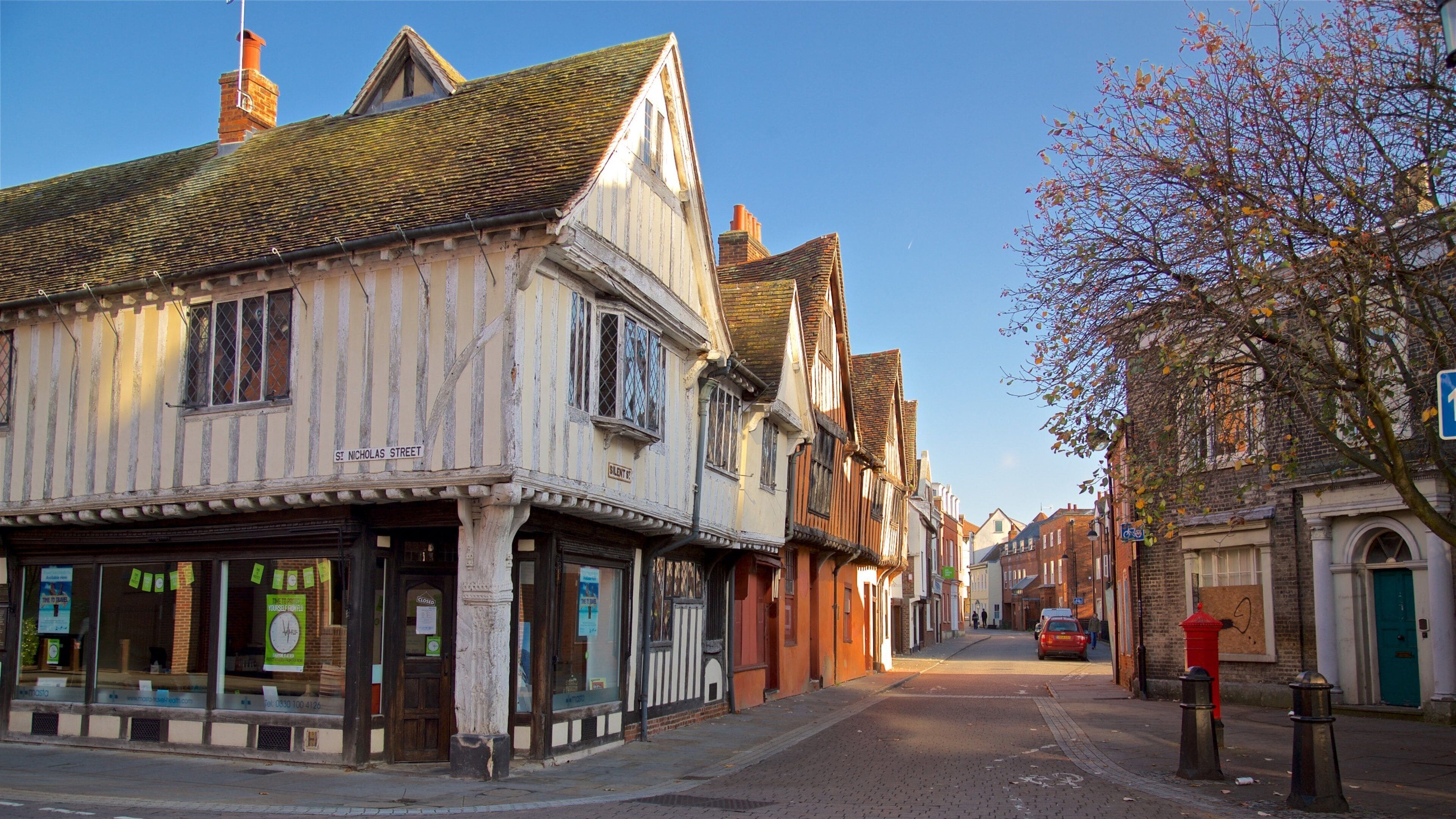 A street lined with historic timber-framed buildings, a red car parked on the side, and a clear blue sky.