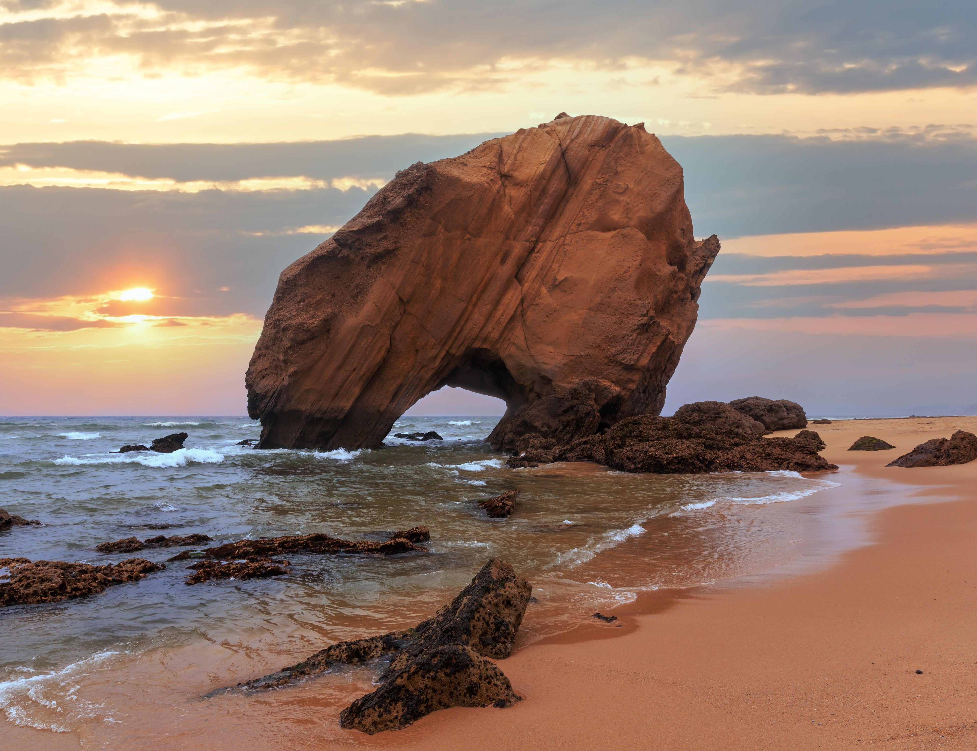 A large rock formation arching over the ocean at sunset.