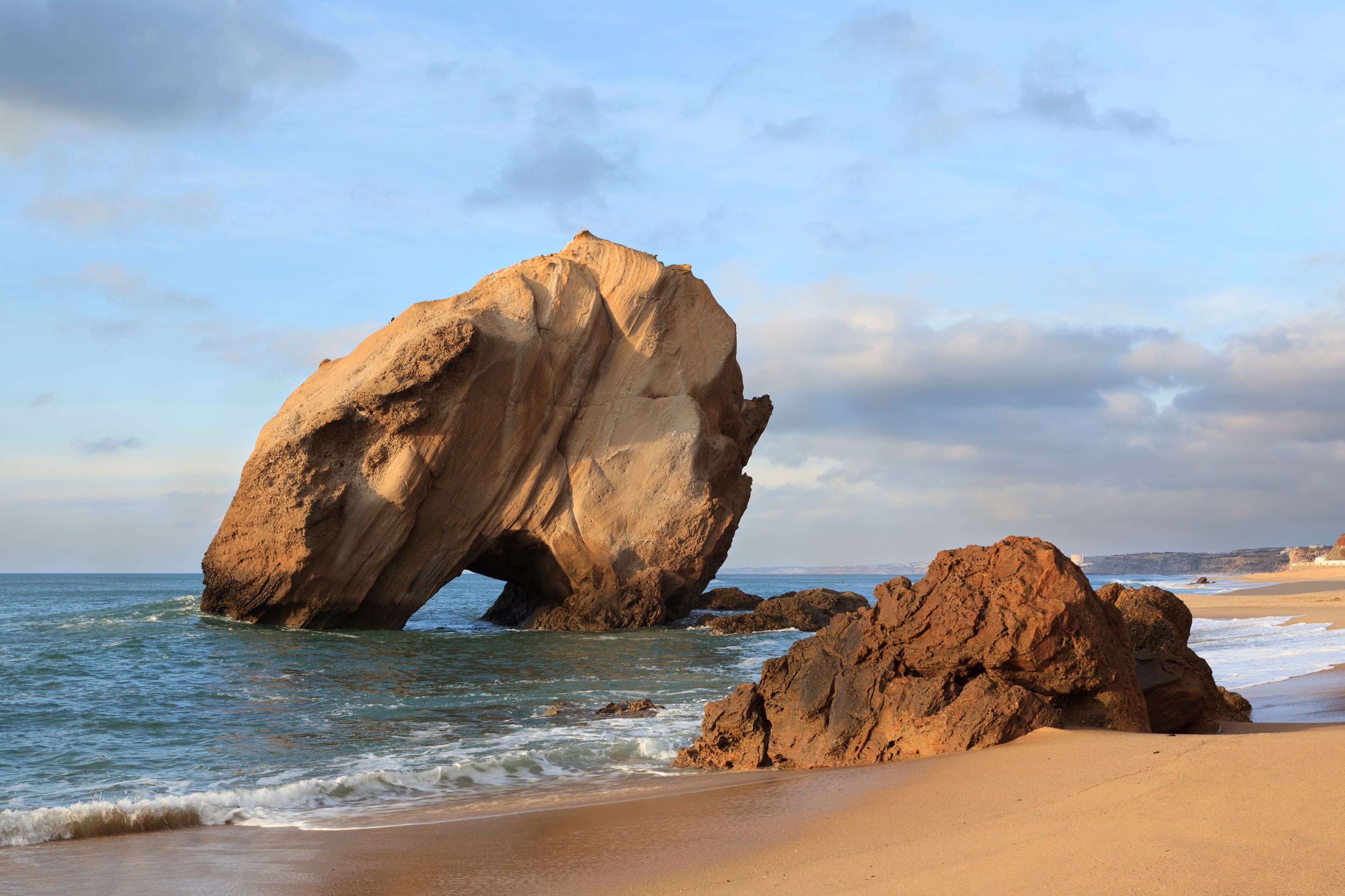 A large rock formation with a natural arch over the sea, a sandy beach, and a clear sky.