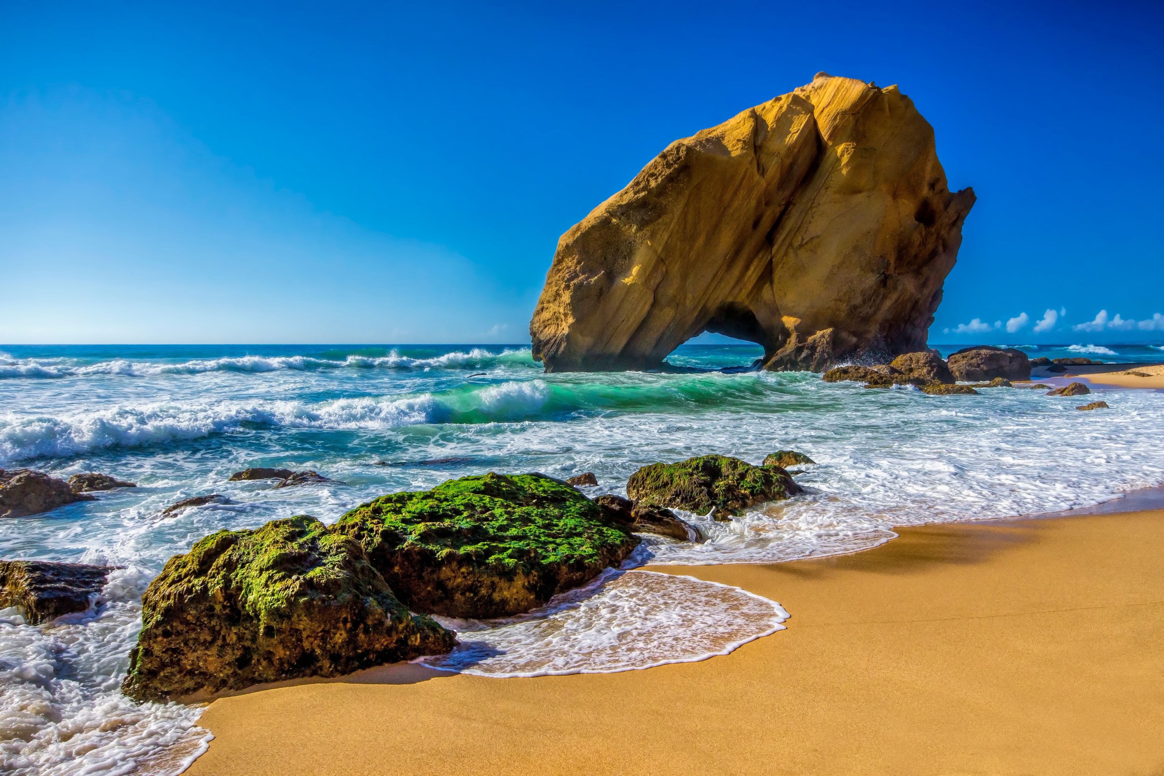 A coastal scene with a prominent rock formation, waves, and a sandy beach.