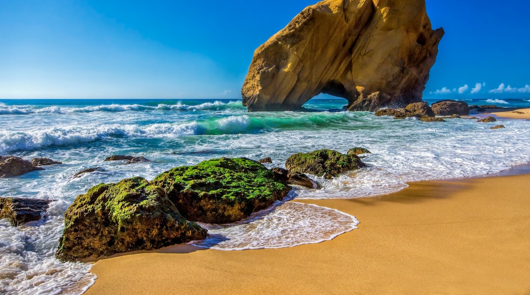 Escena costera con una formación rocosa destacada, olas y una playa arenosa.