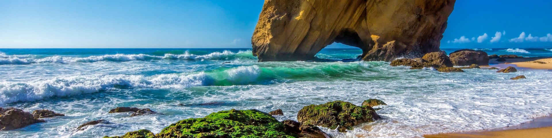 Escena costera con una formación rocosa destacada, olas y una playa arenosa.