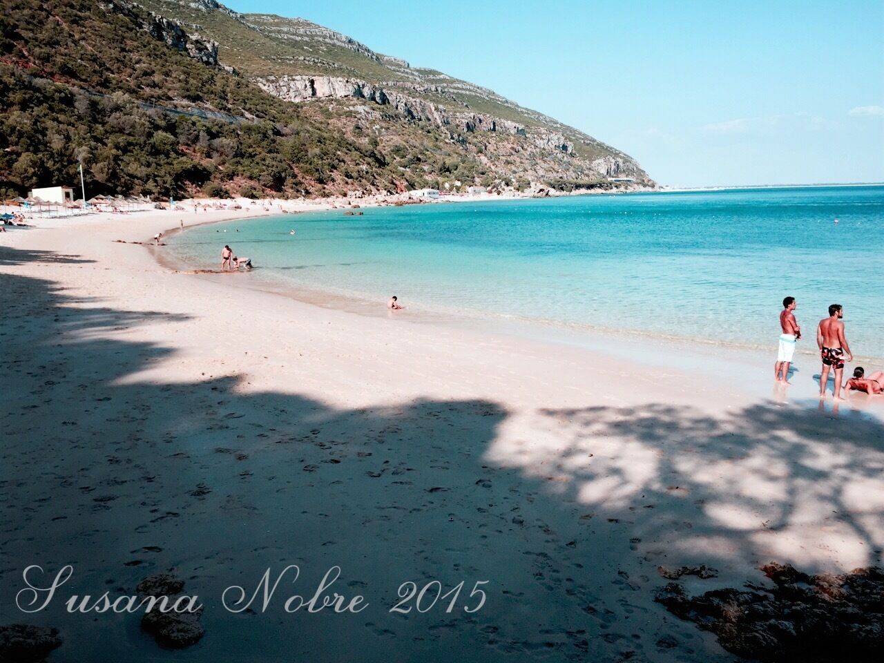 A beach with clear turquoise water, sandy shore, and rocky hills in the background.