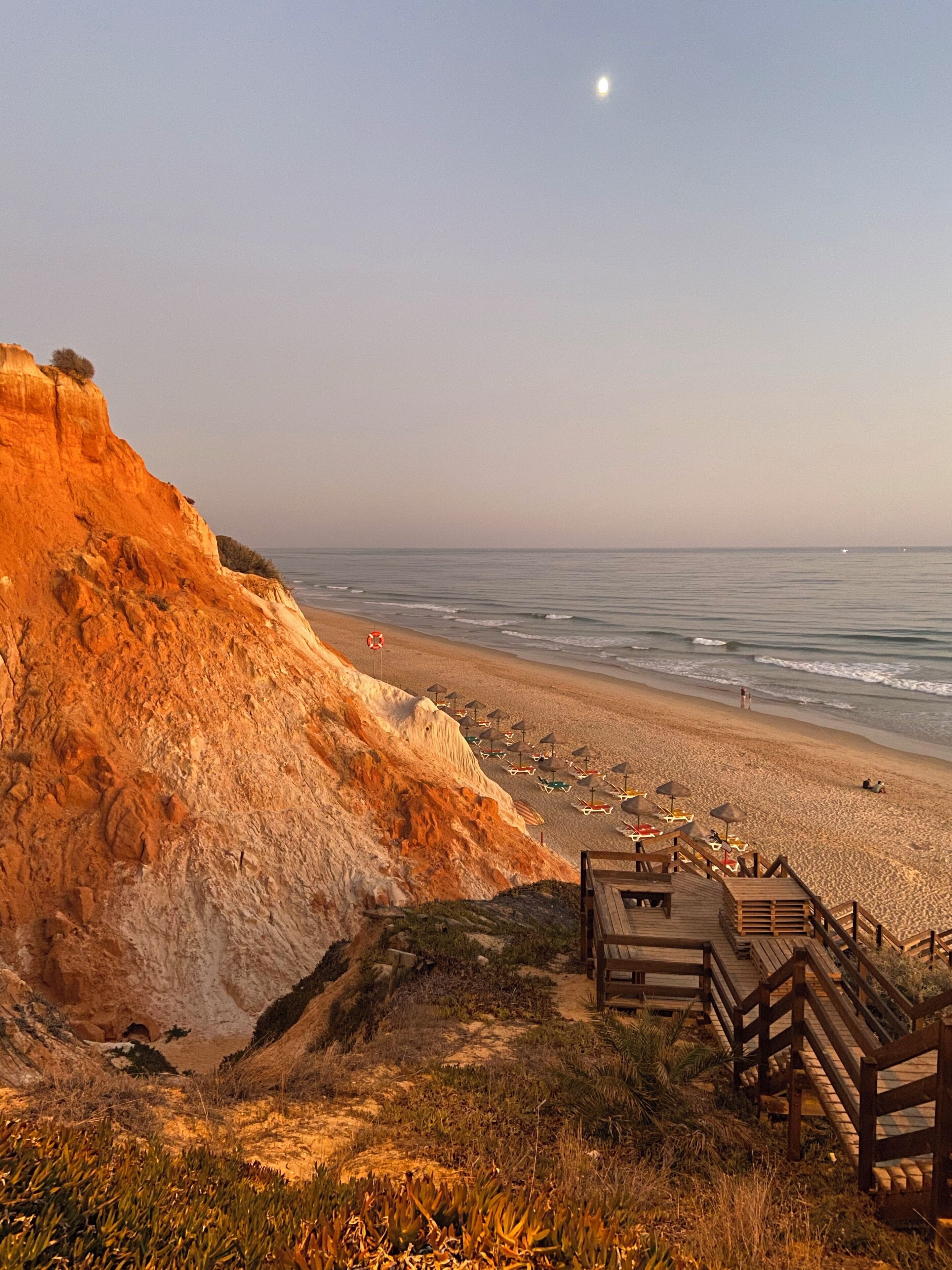 A beach with a wooden boardwalk, umbrellas, and a cliff.