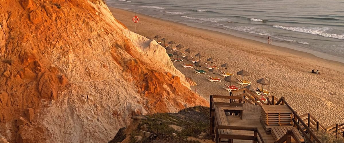 A beach with a wooden boardwalk, umbrellas, and a cliff.