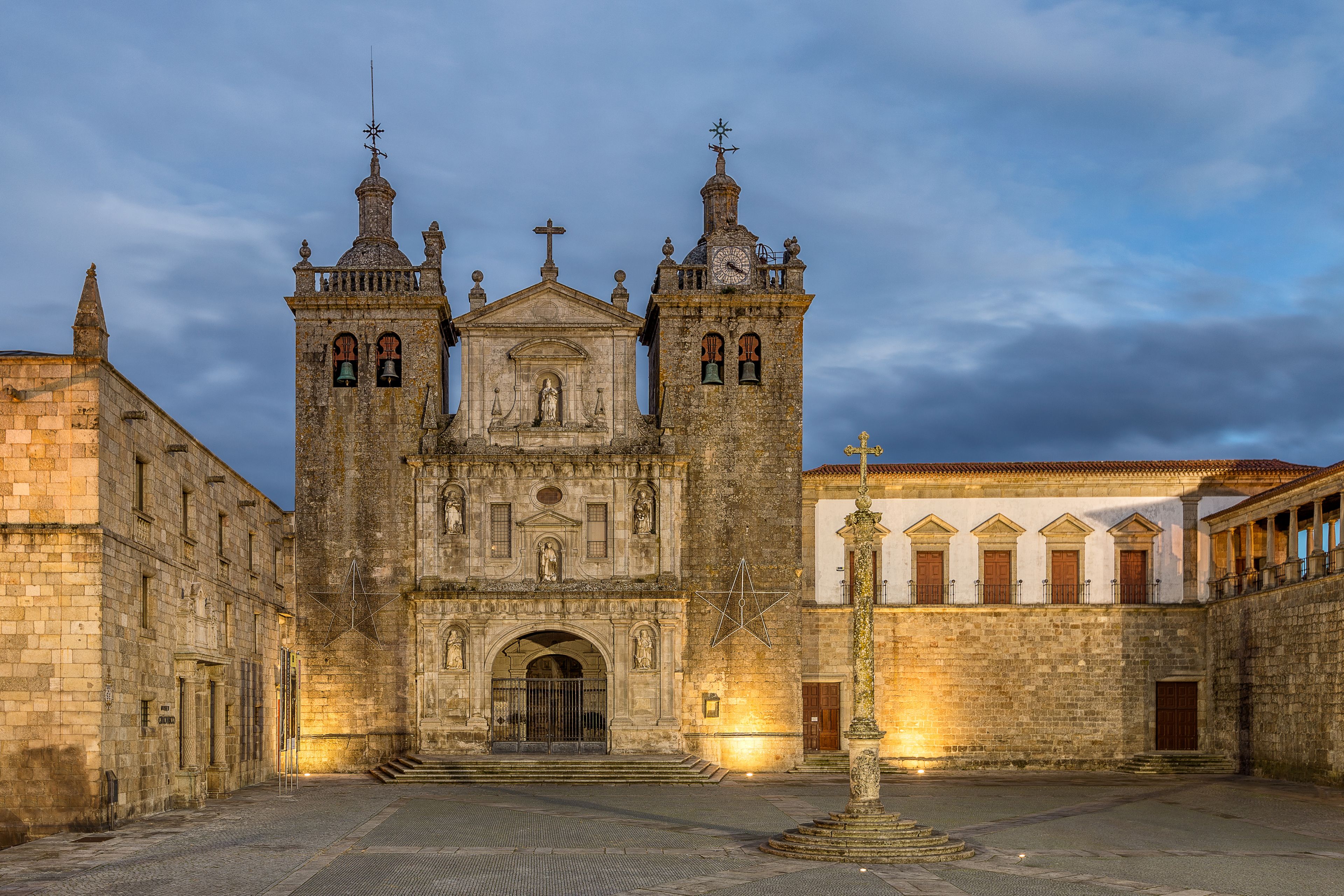 Uma igreja histórica com uma torre central e duas torres menores, rodeada por um pátio com uma fonte.