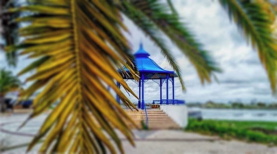 Un quiosco azul con base blanca, rodeado de palmeras y una masa de agua.