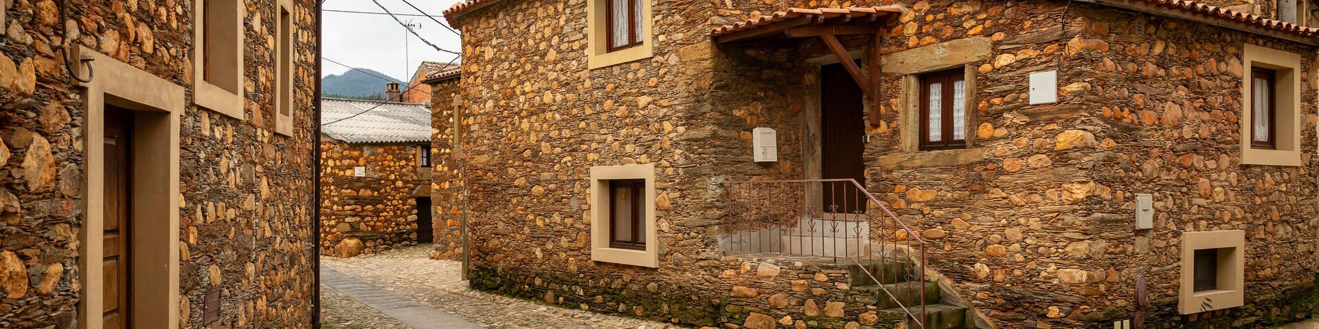 Uma rua de paralelepípedos estreita, ladeada por edifícios de pedra com arquitetura tradicional, incluindo uma escadaria de madeira que leva a uma porta.
