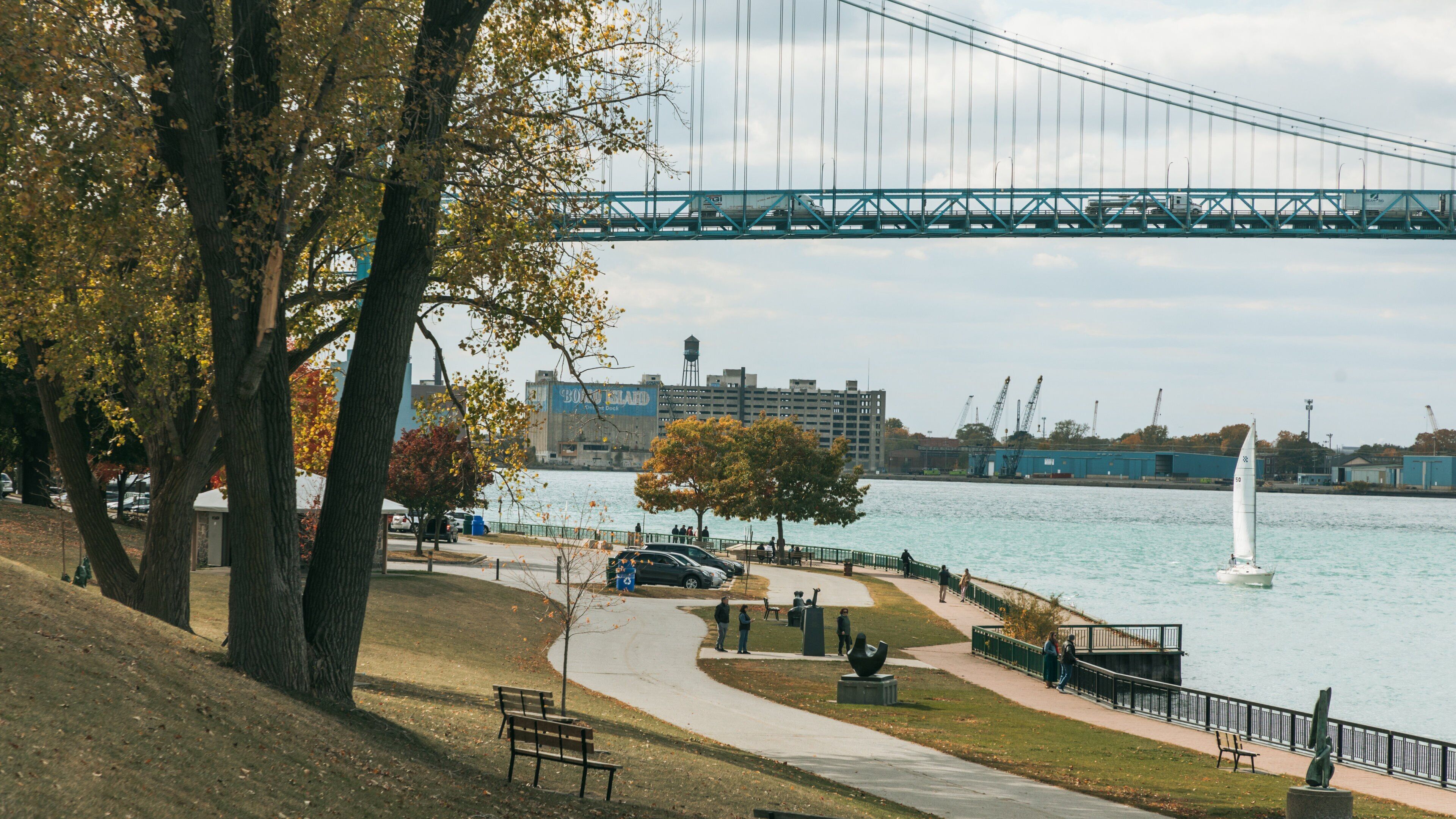 Un parque con un río, un puente y edificios industriales al fondo.