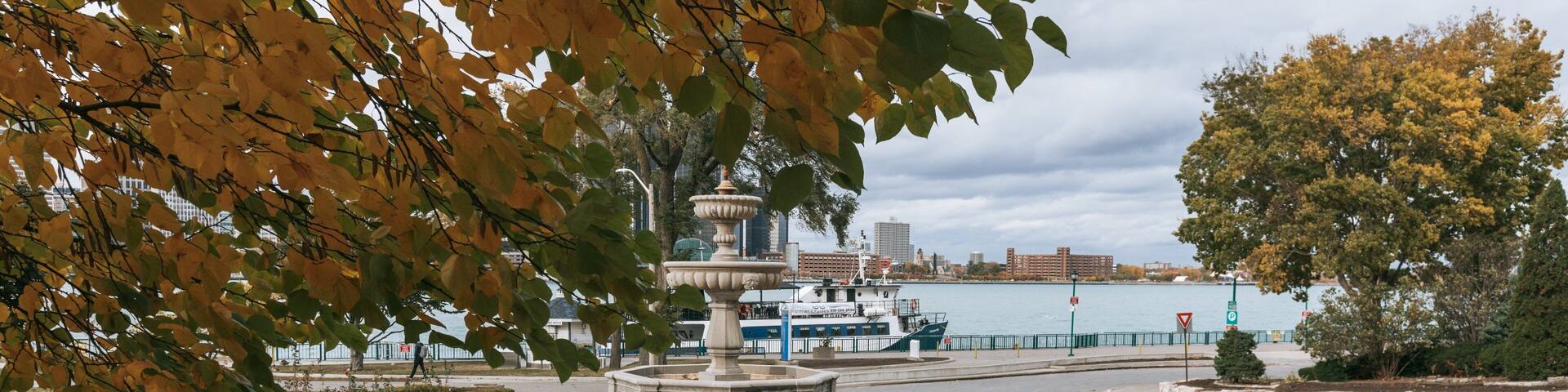 A park with a central fountain, benches, and a body of water in the background.