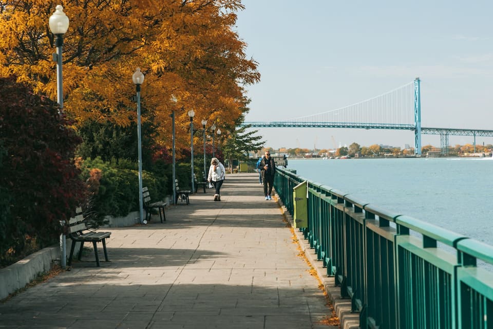 Uma passarela para pedestres ao lado de um rio, com uma ponte ao fundo.