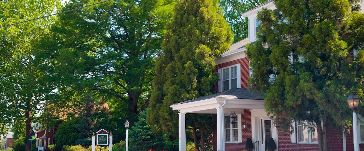 A red brick house with a white porch, surrounded by lush green trees and vibrant red flowers.
