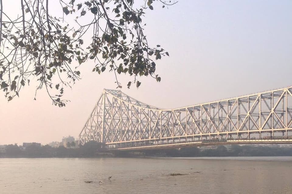 A large steel bridge over a calm river with people sitting and standing on the riverbank.
