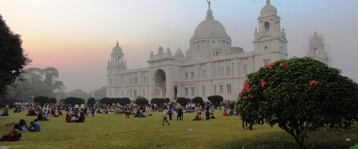 A large white building with domes and towers, surrounded by a grassy area with people sitting and walking.