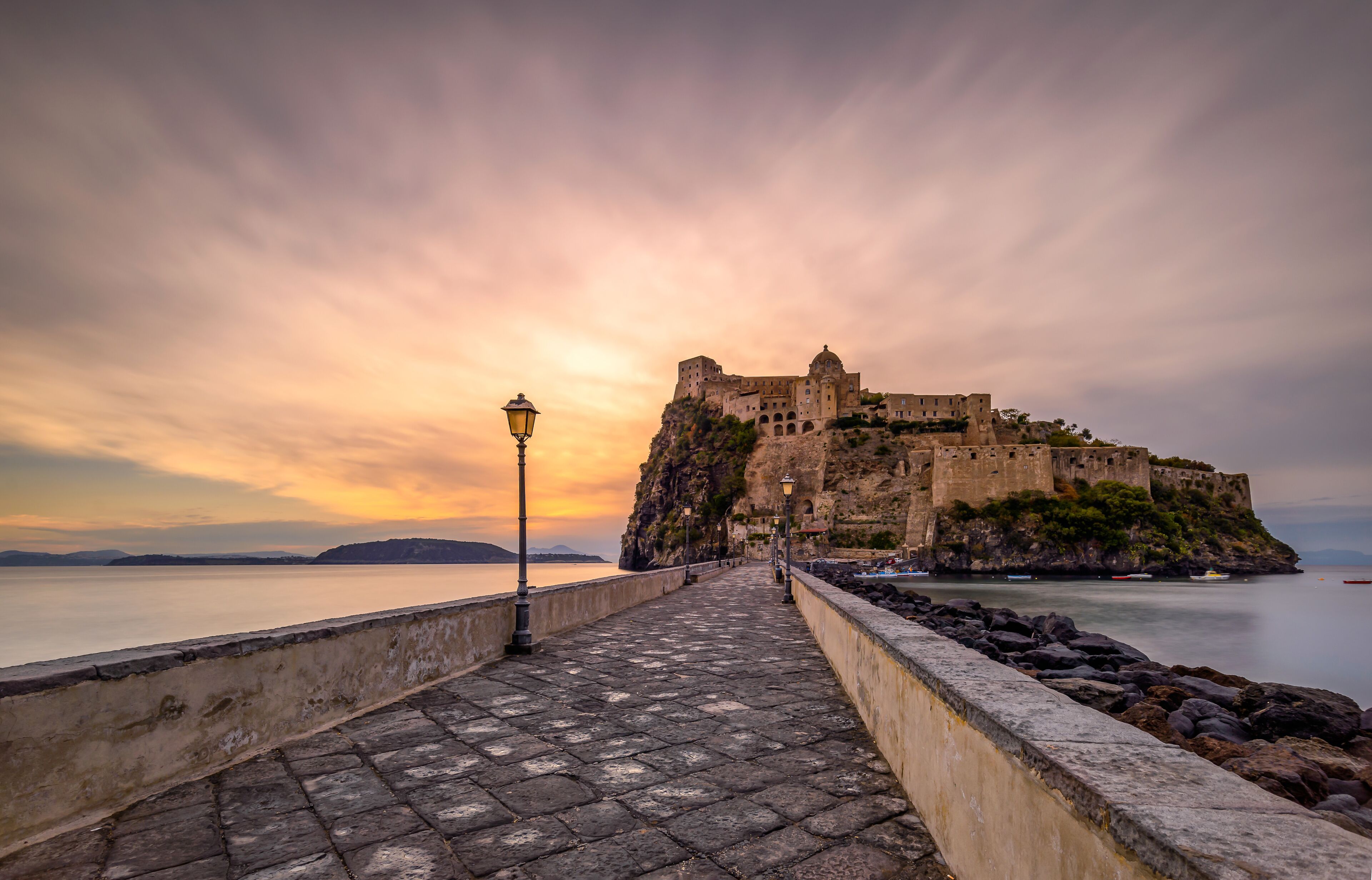 A historic castle on a rocky outcrop overlooking calm waters, with a cobblestone walkway leading to it.