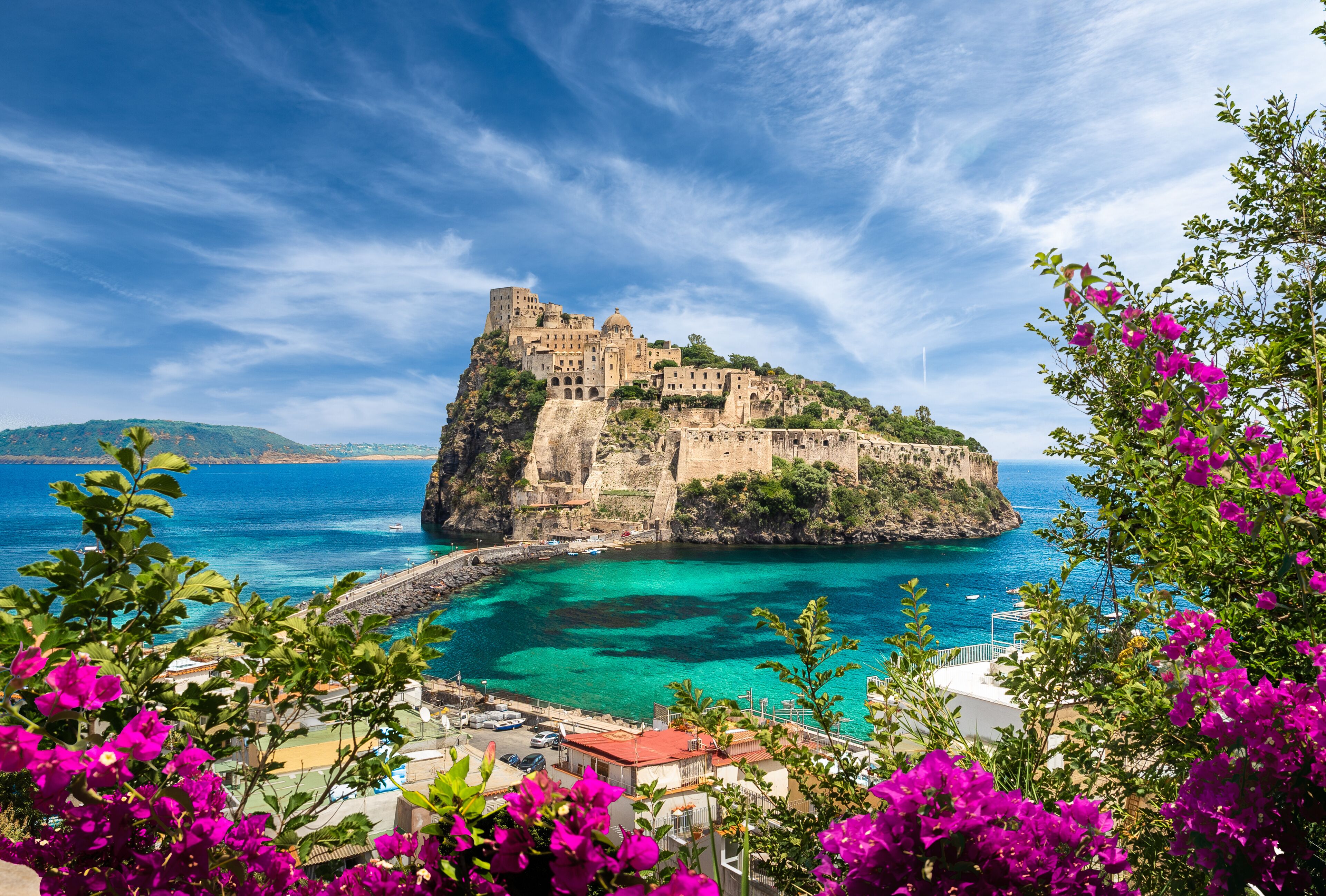 A historic castle on a cliff overlooking turquoise waters, with vibrant bougainvillea in the foreground.
