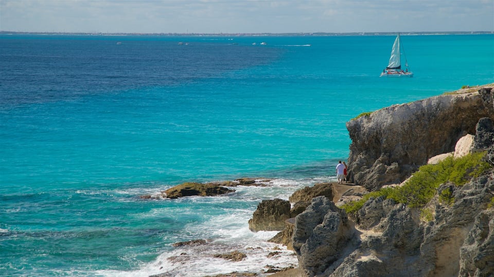 A coastal landscape with clear turquoise waters, rocky cliffs, and a sailboat in the distance.