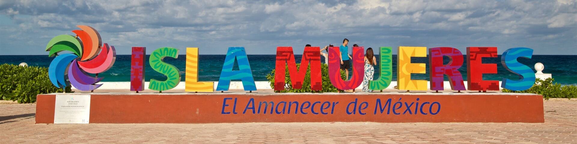 A colorful sign with 'ISLA MUJERES' and 'El Amanecer de Mexico' written on it, set against a backdrop of a clear blue sky and ocean.