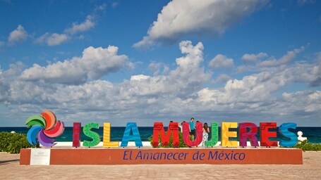 Ein farbenfrohes Schild mit der Aufschrift 'ISLA MUJERES' und 'El Amanecer de Mexico' vor einem klaren, blauen Himmel und dem Meer.