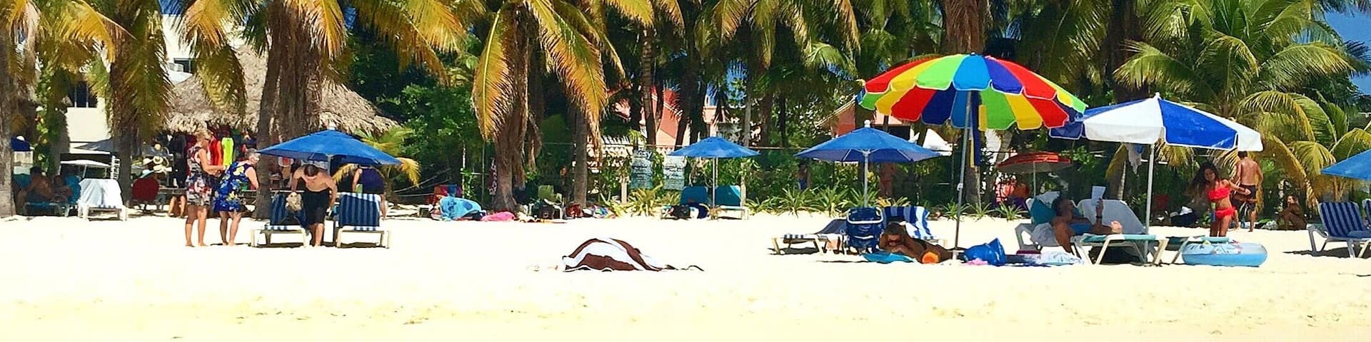 A beach with clear turquoise water, palm trees, and people relaxing under colorful umbrellas.