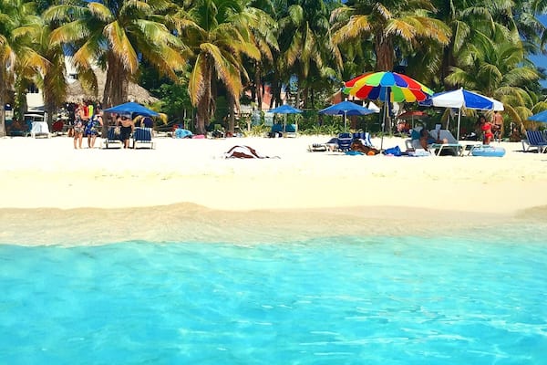 Ein Strand mit klarem türkisblauem Wasser, Palmen und Menschen, die unter bunten Sonnenschirmen entspannen.