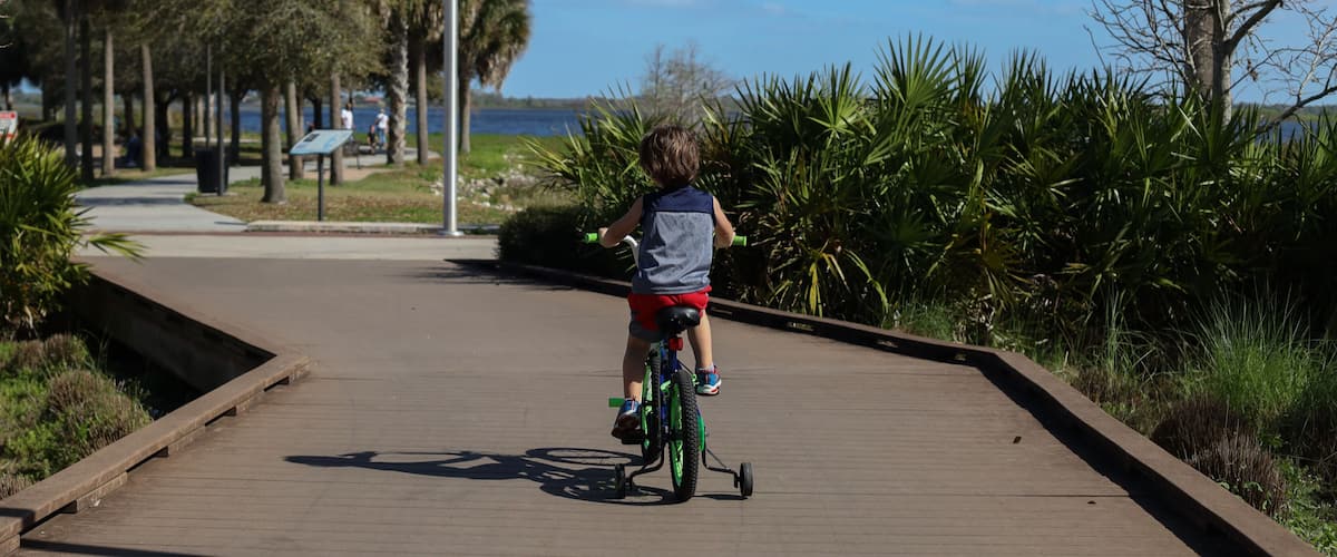 A child riding a bicycle on a wooden boardwalk with a body of water and trees in the background.