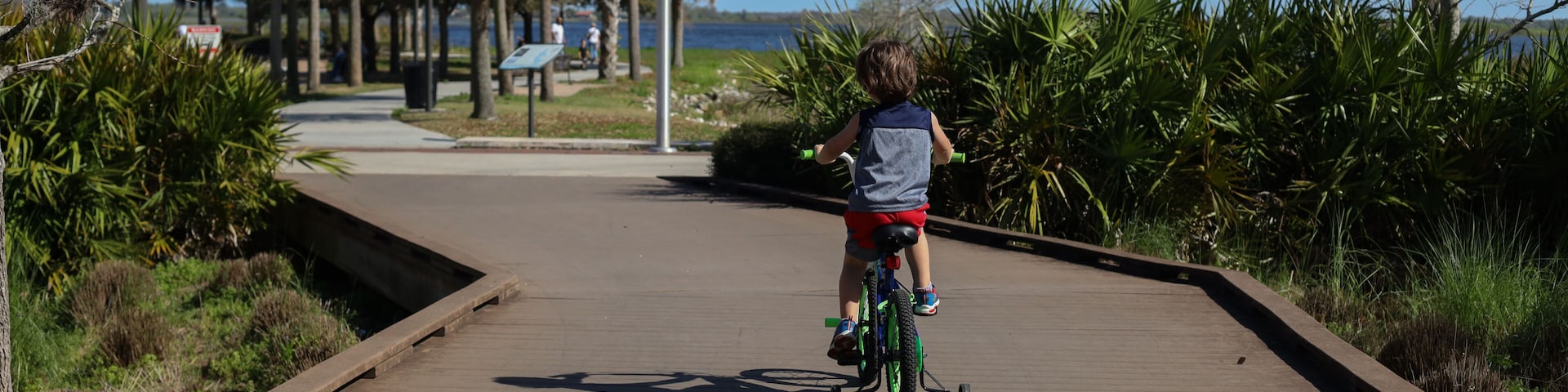 A child riding a bicycle on a wooden boardwalk with a body of water and trees in the background.