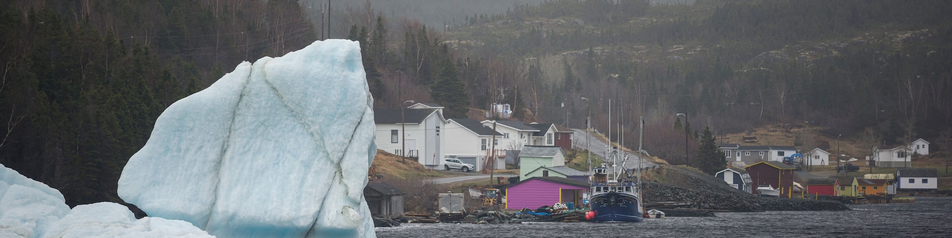 Um icebergue imenso na água, com casas e um barco ao fundo.