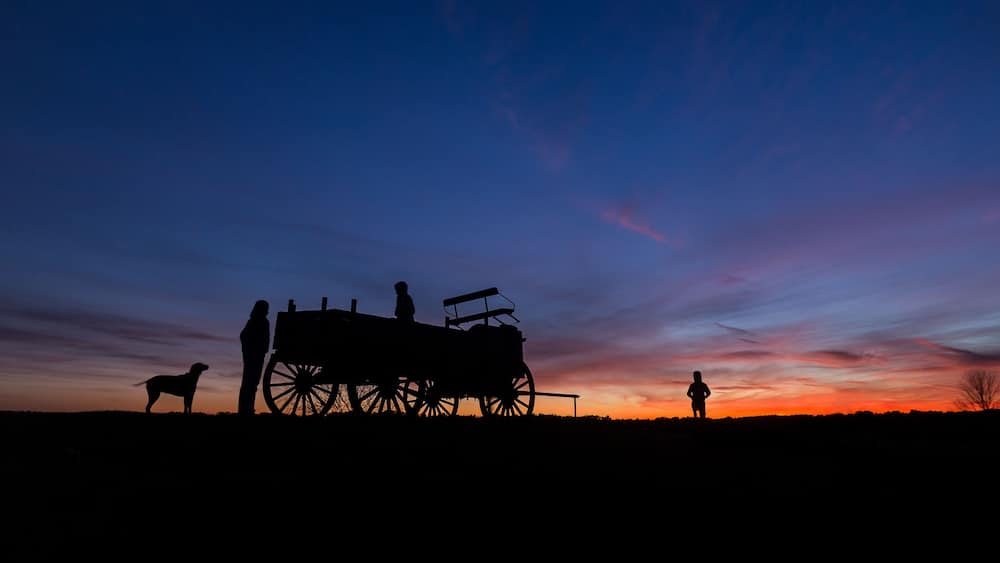 La silhouette d’une personne, d’un chien et d’une calèche tirée par des chevaux, se détachant sur un ciel au coucher du soleil.