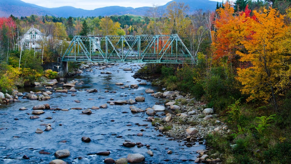 A river with rocks, a bridge, autumn trees, and mountains in the background.