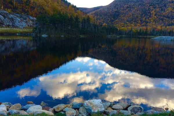 Ein ruhiger See, umgeben von herbstlich gefärbten Bäumen und felsigen Ufern.