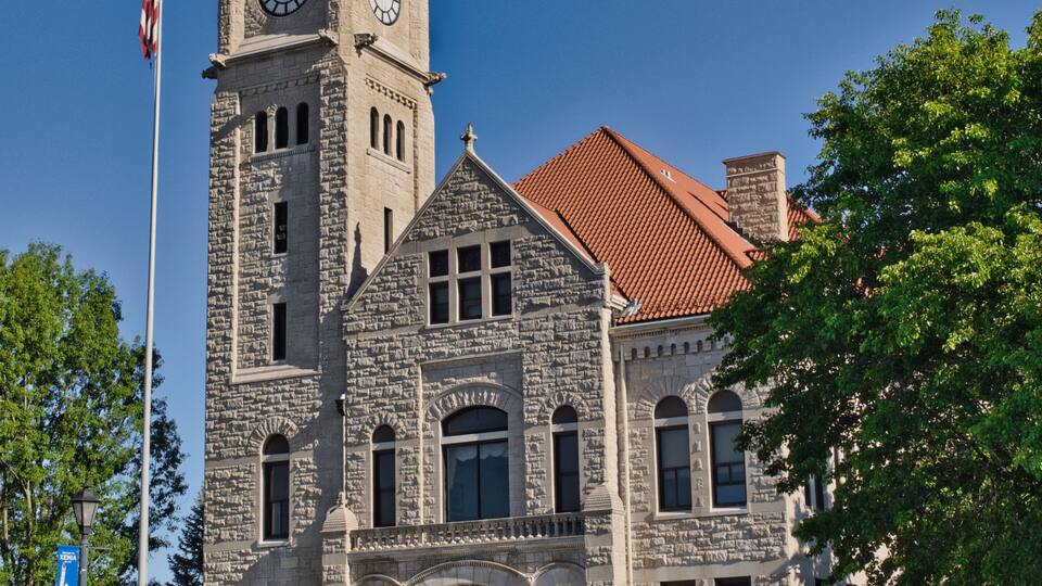 A historic stone building with a clock tower, a red-tiled roof, and an American flag.
