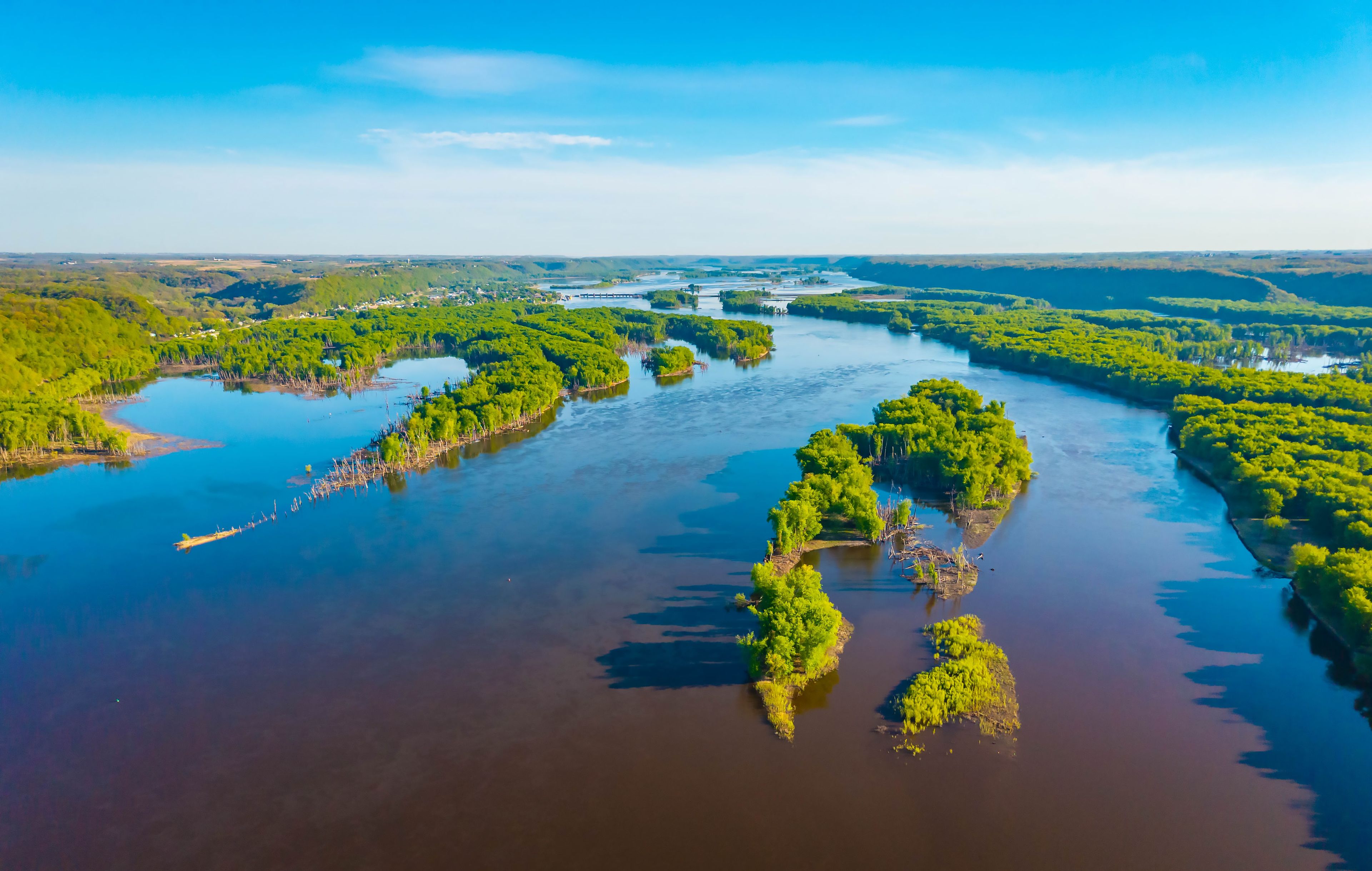 Un río con varias islas cubiertas de vegetación, rodeado por un paisaje con árboles y un cielo despejado.