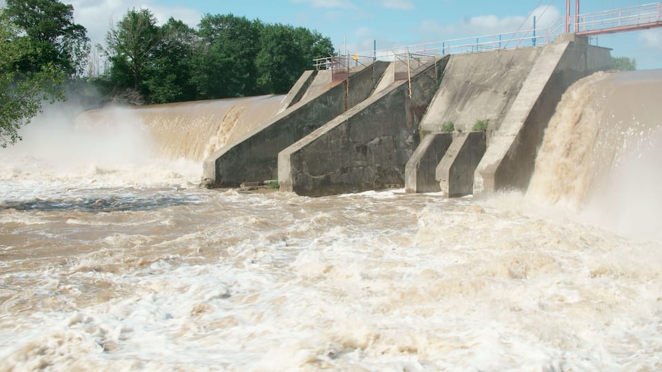 A dam with water cascading over its spillway, surrounded by trees and a clear sky.