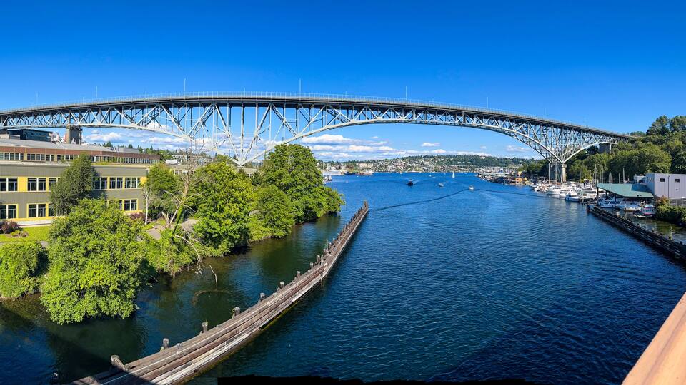 A steel bridge over a river, surrounded by buildings and greenery.