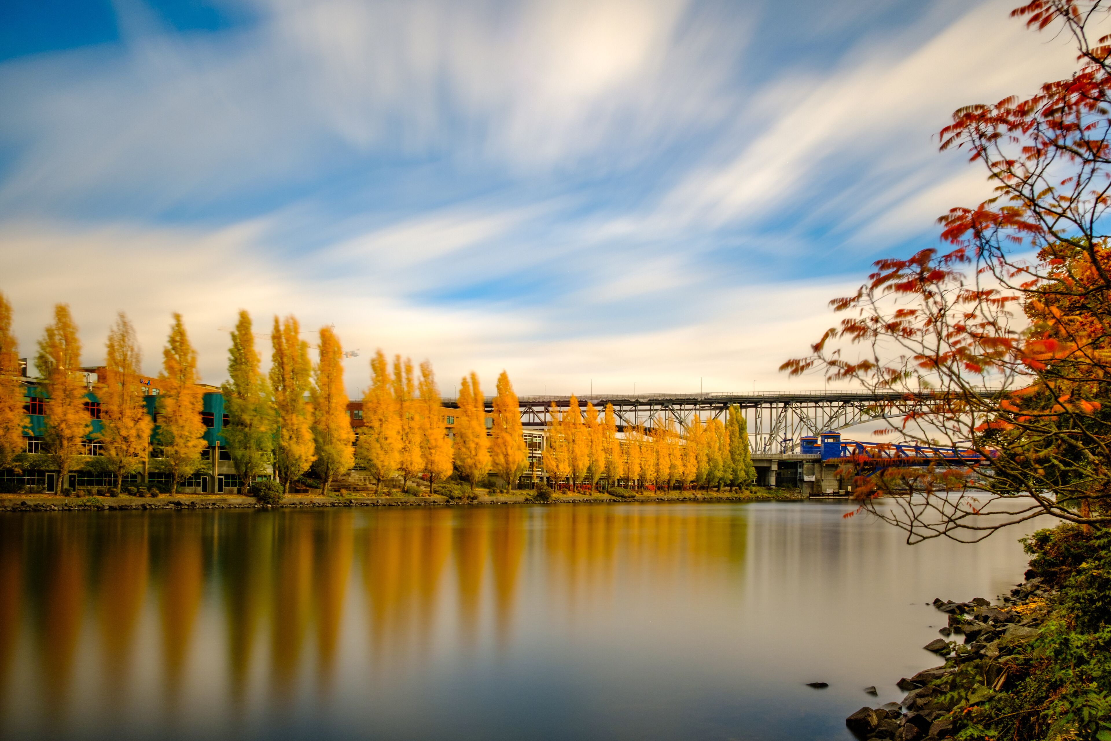 A calm river with autumn trees reflecting in the water, a bridge, and a train passing by.