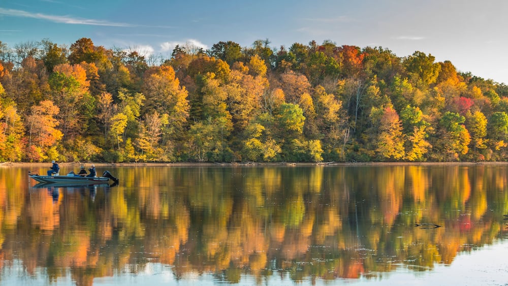 Ein ruhiger See mit Spiegelung von Herbstbäumen und einem kleinen Boot mit Personen.
