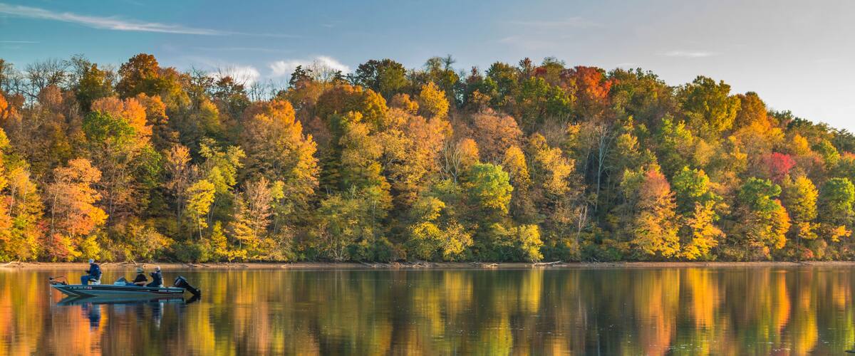 A calm lake with a reflection of autumn trees and a small boat with people.