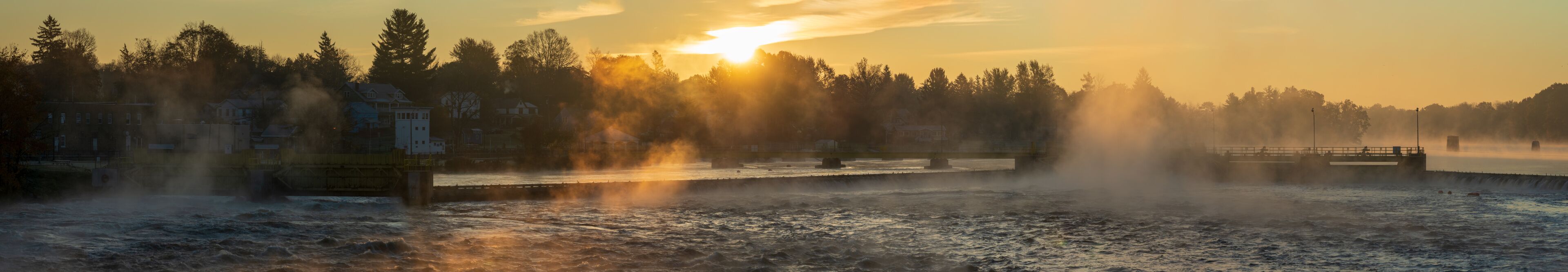 A river with steam rising from the water, a bridge, and a forested shoreline during sunrise or sunset.