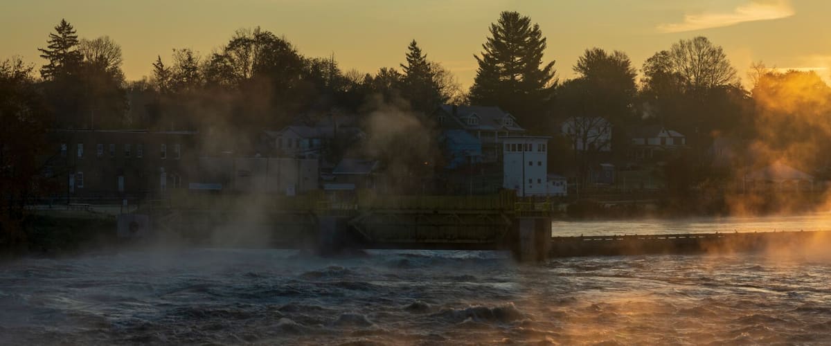 A river with steam rising from the water, a bridge, and a forested shoreline during sunrise or sunset.