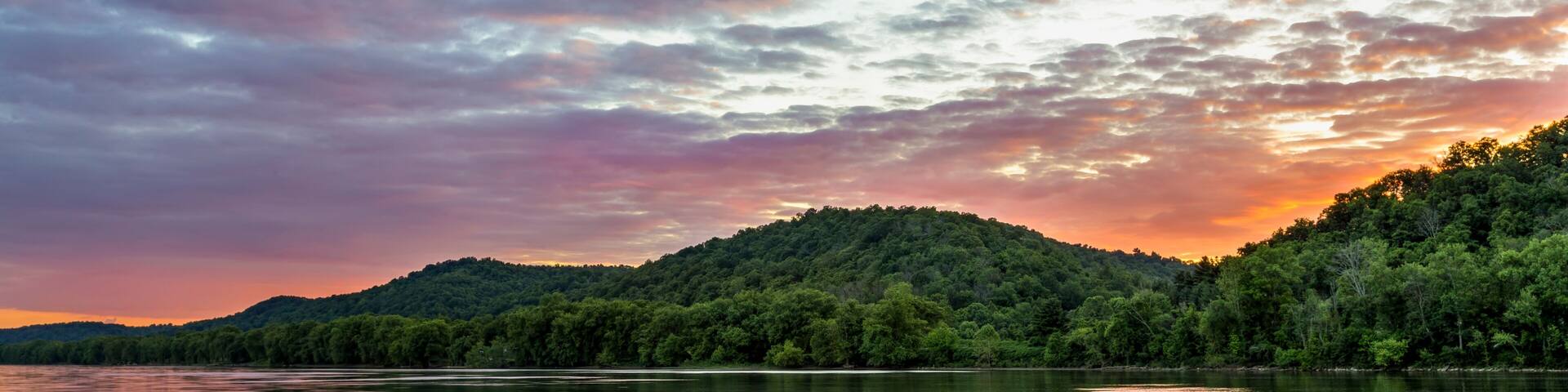 A calm lake with a forested hill in the background and a colorful sky.